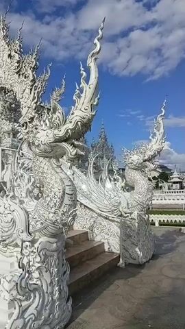 A video shows a close-up view of ornate white structures against a bright blue sky with scattered clouds. The structures appear to be part of a temple or elaborate building, featuring intricate carvings of dragon-like figures and stylized ornamentation. In the foreground, a staircase is flanked by these white sculptures. The camera slowly pans to the right, revealing more of the detailed white architecture and a glimpse of trees and other buildings in the distance. The sunlight highlights the textures and craftsmanship of the white structures, creating strong shadows.