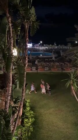 A group of five people are standing on a grassy area at night. They are in a huddle, possibly interacting. In the background, there is a swimming pool area with lounge chairs and umbrellas. Palm trees frame the left side of the scene. The lighting is dim, suggesting it is nighttime.