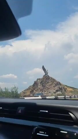 A statue of an eagle with outstretched wings stands atop a rocky, barren hill under a partly cloudy blue sky. The camera is positioned inside a vehicle, looking out through the windshield. The dashboard of the car is visible in the foreground, including a dashboard display screen and air vents. In the middle ground, a highway with two lanes of traffic is visible. Other vehicles, including a white van and a white sedan, are seen on the highway. The landscape is dry and covered with sparse vegetation. The video shows the eagle statue from different angles as the vehicle moves.