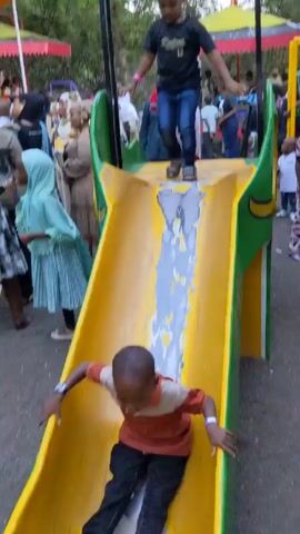 The video shows a group of children taking turns sliding down a yellow and green slide. The slide is located outdoors, seemingly in a park or public area, with a crowd of people watching nearby. The children appear to be enjoying themselves.  The slide shows signs of wear, with some faded or discolored areas in the center. The surrounding environment shows trees and other people, indicating a busy public gathering.