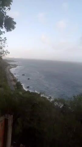 The video pans from left to right, revealing a coastal landscape. In the foreground, foliage and a brick structure are visible. The middle ground features a winding road running alongside a rocky coastline and the ocean. The ocean appears choppy with waves breaking on the shore. In the background, the sky is overcast, with a hazy horizon where the sea meets the sky. The overall impression is of a natural, perhaps remote, coastal environment. There are no people or animals visible in the frame. The lighting suggests it might be early morning or late afternoon due to the diffused light.