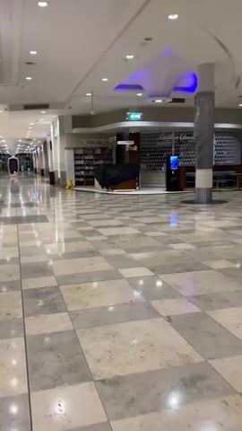 The video shows the inside of a nearly empty shopping mall, looking down a wide corridor with a checkered marble floor and closed storefronts on either side. A signpost with directions to a cinema and customer service is visible. A touchscreen kiosk is present near the signpost. The mall appears deserted with only a few items like trash bins and some covered objects indicating ongoing cleaning or setup. The lighting is bright and even.
