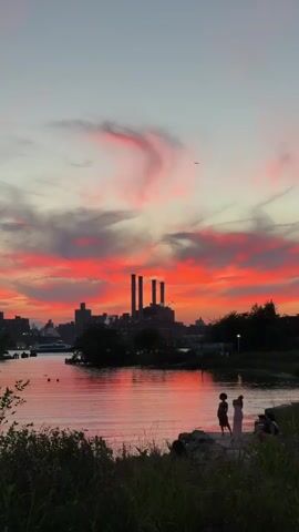 A vertical video captures a sunset over a city skyline. The sky is filled with dramatic clouds painted in vibrant hues of orange, pink, and red, with a hint of gray. In the foreground, a body of water reflects the colorful sky. On the far bank of the water, a cluster of buildings, including a prominent industrial structure with three tall smokestacks, forms the city skyline. Silhouetted against the sunset, on the near bank, are a few people. Two individuals, appearing to be women, stand close to each other, one in a light-colored dress and the other in a darker outfit. Another figure is seated nearby. The immediate foreground is dominated by tall, dark green grass and some rocks. The overall atmosphere is serene and picturesque, highlighting the beauty of the sunset.