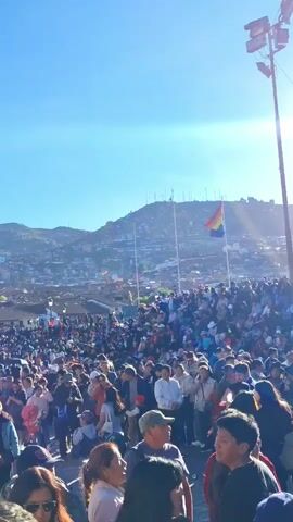 This video captures a lively outdoor gathering in Cusco, Peru. A large crowd of people, diverse in age and attire, fills the frame, suggesting a public event or celebration. Many individuals are holding up their phones, indicating they are taking photos or videos, which is a common behavior at such events. The atmosphere appears festive and engaging, with a sense of collective participation.

In the background, a hilly landscape dotted with buildings suggests an urban setting, likely Cusco given the context. A prominent rainbow flag is visible, waving from a pole, which could signify a pride event or a celebration of diversity. The sky is a clear, bright blue, and the bright lighting indicates it is daytime, likely mid-morning or afternoon. The sun's position and the general brightness suggest fair weather.

Among the crowd, several objects are noticeable. Besides the ubiquitous mobile phones, there are hats worn by some individuals, likely for sun protection. The architecture in the background appears to be traditional, with terracotta roofs, fitting for a historical city like Cusco. The overall impression is that of a vibrant community event, possibly a parade, festival, or demonstration, taking place in a scenic and populated urban environment. The energy of the crowd, combined with the bright daylight and clear skies, points to an enjoyable and possibly significant occasion for the attendees.
