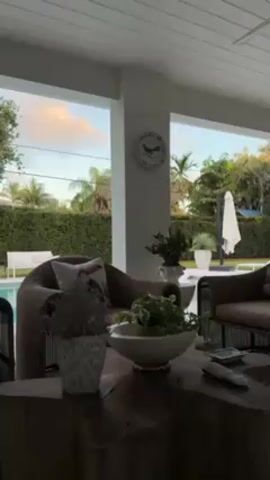 A still shot displays an outdoor patio area. In the foreground, a dark wooden table with potted plants sits beneath a white ceiling. Beyond, two brown wicker chairs flank the table, each adorned with decorative pillows. Through the white pillars of the patio, a backyard scene unfolds: a swimming pool, white lounge chairs, and a closed white umbrella, all enclosed by a tall, lush green hedge. The sky above reveals a gradient of blue and orange hues, indicative of either dawn or dusk. A decorative plate hangs on one of the white pillars.