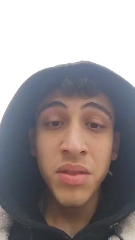A young man wearing a black hooded jacket stands outdoors. The camera focuses on his face, particularly his mouth and nose. He is speaking. A road and a tree are visible in the background. The sky is overcast.