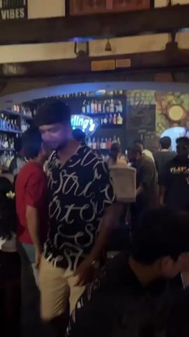 The video shows the interior of a bar, labeled "Darling's Bar" with a blue neon sign. The bar area is well-stocked with bottles. Numerous people, mostly men, are gathered around the bar. Some patrons are seated at tables, while others stand near the bar, talking. The bar has a rustic ambiance, with wooden beams, paintings, and decorative signs. The lighting is relatively dim, contributing to the bar's atmosphere. The camera pans slowly across the room, showing the layout and crowd.