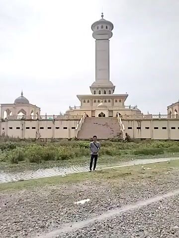 A man walks on a gravel path towards the camera, with a large, elaborate architectural complex in the background. The prominent building features a tall, cylindrical minaret with a domed top, situated atop a multi-tiered base. Two smaller, domed structures flank the central building, connected by lower walls that define a broad, elevated platform accessible by a wide staircase. The building is light-colored, possibly cream or beige.

In front of the building, a wide area of sparse grass and dirt separates the man from the structure, with a narrow, muddy-looking ditch or waterway running horizontally across the midground. The ground where the man stands is covered in gravel and small stones. The man wears a light-colored short-sleeved shirt, dark trousers, and a dark bag slung across his chest. He walks forward, looking towards the camera, with a slight smile. The scene is brightly lit under a clear, overcast sky, indicating daytime. The architecture, characterized by domes and a minaret, strongly suggests an Islamic religious or cultural site, consistent with the location of Kruengmate, Indonesia. There is no visible text in the image.