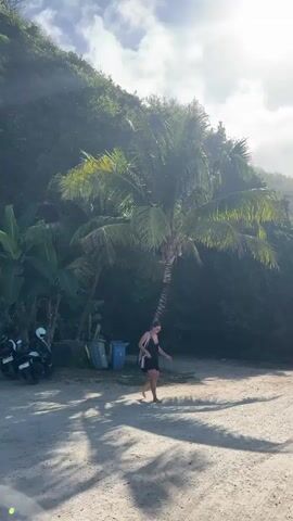 A woman walks across a sandy area in front of a lush green hillside under a partly cloudy sky. The sun is shining brightly through the clouds, casting strong shadows. The woman is wearing a black dress and carrying a striped bag over her shoulder. A palm tree stands to her left, and a couple of trash bins are visible behind it. A motorcycle is parked to the far left of the frame. A small building with a light-colored exterior is visible in the background on the right. The woman is walking away from the camera, toward the right side of the frame.
