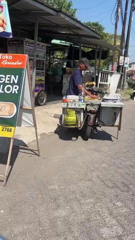 The image shows a street vendor in Kalisampurno, Indonesia, preparing food from a mobile cart. The vendor, a man wearing a blue shirt and a black cap, is focused on his work. The cart is a repurposed motorcycle with a food preparation station attached. A green gas cylinder is prominently displayed on the cart, likely powering a cooking appliance. In the background, other stalls and shops are visible, suggesting a busy marketplace or street. A white easel sign in the foreground displays text in Indonesian, including "Toko," "Karisendra," and other phrases that are partially obscured. The sunny weather and clear sky indicate it is daytime. The overall scene depicts a typical street-food vending operation in an urban or semi-urban Indonesian setting.