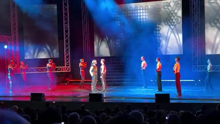 Six men are on a stage. Two men in the center of the stage stand facing each other. Both men are wearing costumes that appear to be elaborate and ornate, with a mix of dark and metallic colors. The man on the left wears a helmet with a visor and a chest plate with intricate designs. He also wears dark pants with red accents and what appear to be shin guards. The man on the right wears a sleeveless top with decorative detailing on the chest and dark pants.

To the left of these two men, three other men are positioned. They are all dressed in dark, fitted outfits with red accents. Two of them are wearing helmets with visors. The third man, standing slightly apart, has his hands on his hips and appears to be watching the central figures.

To the right of the central figures, three more men are positioned. They are all wearing sleeveless tops and dark pants. Their chests are bare, and they appear to be in various stances, possibly posed or engaged in light movement.

The stage is lit with a combination of red and blue lights, and there is smoke or haze filling the air, creating a dramatic atmosphere. Behind the performers, there is a backdrop that appears to be a large screen displaying abstract patterns or imagery. A set of tiered platforms or stairs is visible behind the central figures. In the foreground, the audience is visible, seated in a dimly lit theater. The overall scene suggests a performance, possibly a dance or theatrical show, with a strong emphasis on costumes and dramatic lighting.