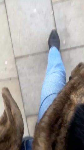 The video shows a first-person view of someone walking on a paved sidewalk. The person is wearing blue jeans and black pointed shoes. A brown furry jacket is visible on either side of the frame. The camera angle is pointed downwards, capturing the person's legs and the ground they are walking on. The sidewalk is made of grey and beige rectangular paving stones. The person is walking at a moderate pace.