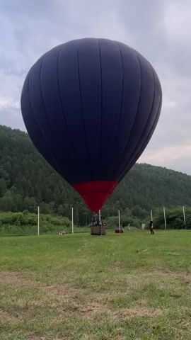 A hot air balloon is being prepared for flight in a grassy field. The balloon is dark blue with a red bottom and is attached to a basket where two people are visible. The balloon is tethered to the ground by white poles. In the background, there is a wooded hill and a cloudy sky. Further in the distance, there are people and a car, and a stage with red fabric draped from a central pole. The overall scene suggests an outdoor event or festival.