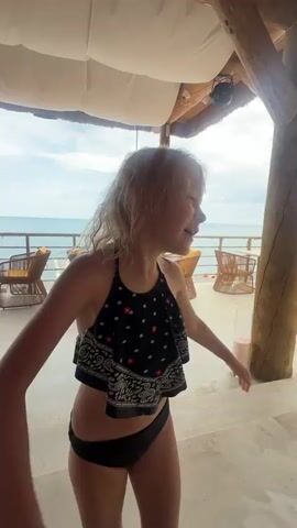 A young girl with blonde hair, wearing a black bandana-style bikini top and black bottoms, poses in an outdoor setting. She is initially seen in profile, looking towards the ocean, then turns to face the camera with a wide, playful smile, revealing missing front teeth. Her hair is wet and styled loosely.

The background suggests a beachside establishment with a thatched roof supported by rustic wooden beams. White fabric drapes hang from the ceiling, and a light fixture is visible. In the mid-ground, wicker chairs with yellow cushions are arranged on a concrete patio. Beyond the patio, a railing overlooks the ocean. The sea is calm, and the sky is a clear blue with some clouds. Sunlight filters through the structure, indicating it is daytime, likely mid-morning or afternoon. A tall, pink candle in a glass holder sits on a table to the right of the frame.

The girl's expressions shift from a thoughtful gaze to an enthusiastic and direct engagement with the camera, conveying a sense of fun and mischief. The overall atmosphere is relaxed and vacation-like, consistent with a seaside resort. No visible text is present in the media.