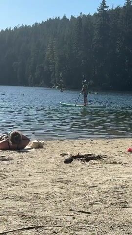 The video shows a sandy beach next to a lake. A person is lying on the beach with their back to the camera. Further out on the lake, a person is paddleboarding. The background is a forest. The sky is clear and blue. The water is calm with ripples.