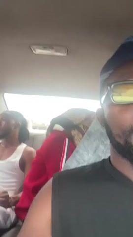 The video shows the interior of a car, with three people visible. The primary subject is a young Black man in the front passenger seat, wearing a black tank top and sunglasses. He is speaking directly to the camera, gesturing with his hands. In the backseat, a young Black man with braids and a white tank top is seated on the left, looking forward. Next to him, another person is wearing a red hooded jacket and a patterned headscarf, obscuring their face. The scene appears to be outdoors, with glimpses of greenery and a cloudy sky visible through the car windows, suggesting it is daytime. The car's interior is simple, with light-colored seats and a ceiling light. The overall mood seems casual, possibly a road trip or a ride. No visible text is present in the media. The location context provided is Kwali, Nigeria.