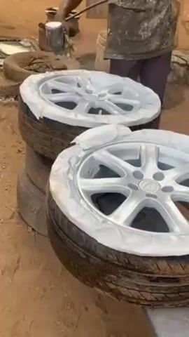 In an outdoor setting, a person is seen spray painting car wheels that are stacked on tires. The individual, likely a painter, is wearing dark pants and a light-colored shirt covered with paint splatters. They hold a spray gun and apply a light-colored paint to the wheels. The wheels are partially covered with tape, presumably to protect the tires from the paint. The wheels are sitting on stacks of tires on the ground. The surrounding area appears to be a workshop or outdoor workspace, with other tires, tools, and a bucket visible in the background. The ground is dirt.