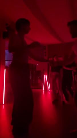 In a dimly lit room bathed in red light, two silhouetted figures dance facing each other. The first, presumed female, wears a crop top and a long skirt. The second figure, presumed male, is in a light-colored t-shirt and dark shorts. Red LED strips illuminate the floor, adding to the red ambiance. Reflections in the mirror reveal the presence of other people in the background. A tripod stand sits near the back of the room. The red lighting and motion blur make it difficult to see details.