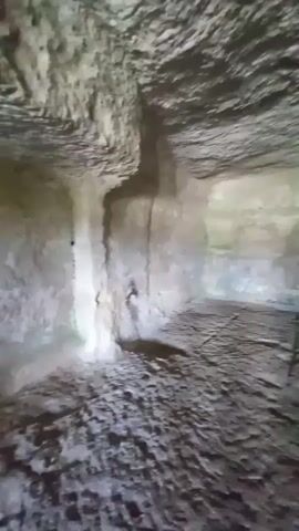 The video pans across the interior of a rough-hewn cave or underground chamber. The walls, ceiling, and floor appear to be made of rock, with an uneven, textured surface. There is evidence of carved-out niches or shelves in the walls, suggesting it may have been used as a dwelling or storage space. The lighting is dim, with natural light entering from an opening at the end of the chamber, revealing a set of stone steps leading upwards. The floor is covered in debris and loose stones. The overall impression is of an ancient, possibly abandoned, subterranean space.