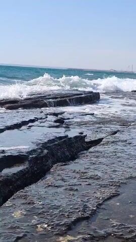 The video captures a dynamic oceanfront scene under a clear sky. The foreground is dominated by textured, dark rocks, some of which are submerged in shallow pools of water, reflecting the light and creating shimmering surfaces. Waves crash against these rocks, generating white foam that drifts across the wet stone. The water itself appears choppy and turquoise, suggesting a lively sea. In the distance, beyond the rocky shoreline, a hazy horizon is visible. There are some indistinct structures on the horizon, possibly industrial or architectural, and a hint of a beach or landmass further away. A few small figures can be seen in the distance along the coastline, suggesting human presence. The bright sunlight and clear sky indicate a daytime setting, likely pleasant weather. The overall impression is one of natural beauty and the raw power of the sea meeting the land.