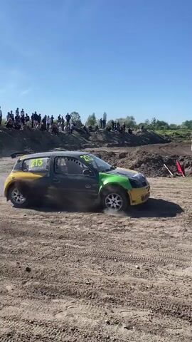 The video shows a small racing car spinning its wheels and kicking up dust as it pulls away from the start line of a dirt track race. The car, number 213, is painted in a mismatched color scheme of yellow, gray, and green. A man stands near the start line, waving a green flag. Spectators stand atop a dirt embankment in the background. The sky is clear and blue.