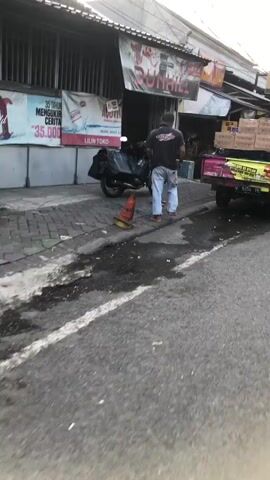 The media file shows an outdoor scene in what appears to be a commercial area, possibly a street market or a row of shops. Two individuals are the primary subjects, engaged in an interaction. One person, wearing a dark t-shirt and jeans, is walking away from the camera, while another, in a lighter-colored shirt, is facing them. They seem to be in a conversational or possibly a dispute.

In the background, there are several elements that provide context. A motorcycle is parked on the sidewalk, with bags on its rear. A truck with boxes stacked in its bed is also visible, suggesting commercial activity or delivery. A shopfront with signage is present. One sign prominently displays "DUNHILL" in large letters, and another, partially obscured, shows "AQUVIA" with text below it. There's also a sign with Indonesian text that reads "LILIN TOKO MURAH SEHAT SELALU".

The setting appears to be a paved street with a sidewalk. There's some debris or water on the road surface, and a traffic cone is placed on the sidewalk. The overall lighting suggests it might be daytime, possibly late morning or afternoon. The mood is neutral, with no clear indications of strong emotions. The location cues, such as the Indonesian text on the signs, strongly suggest the scene is in Indonesia, specifically Ngaban, East Java, as provided.