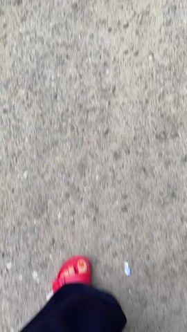 A person's feet are shown from a top-down perspective, walking on a coarse, gray, gravelly surface. The person is wearing bright pink sandals with orange-painted toenails and dark blue trousers. The camera is moving, suggesting the person is walking. The lighting appears to be natural daylight.