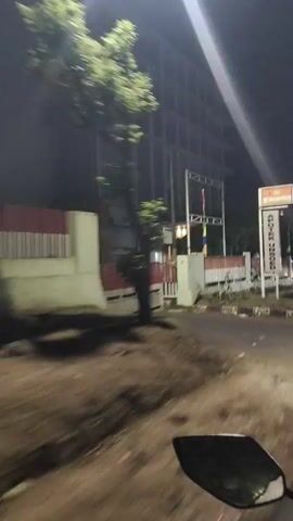 The video shows a nighttime exterior view of a multi-story building with a sign that reads "HOTEL LANMODO." The building is tall and appears to have many floors and windows. A fence with red and white accents runs along the front of the property. There are some trees and foliage visible. The footage appears to be taken from a moving vehicle, as indicated by the motion blur and the partial view of a vehicle's side mirror or bodywork in the foreground of the first frame. The lighting is dim, typical of nighttime conditions, with artificial lights illuminating the building and its signage. Flags are also visible on poles in front of the building. The overall impression is that of a hotel or a similar establishment seen from a street at night.