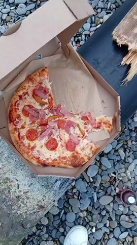 The video shows a pizza in a cardboard box on a rocky beach. Several slices are missing from the pizza. A black pipe and a piece of driftwood are also visible near the box. The camera then pans to show the rocky beach and the ocean waves. The sky is overcast with a hint of sunlight on the horizon.