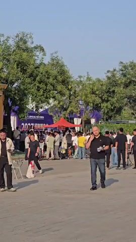 A group of people are gathered outdoors. In the foreground, a bald man in a black polo shirt and blue jeans walks towards the right of the frame, holding a bottle and a phone to his ear. To his left, a man in a light-colored jacket and dark pants walks towards the left, carrying a white shopping bag. Behind them, a crowd of people are congregated around tents and stalls set up outdoors. Trees with green foliage are visible in the background, and the sky is clear and blue. Some of the tents have banners with Chinese text. The ground appears to be paved.