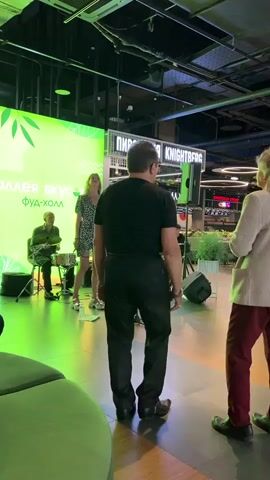 In a brightly lit indoor space with green lighting, a live band performs on a stage. A female singer in a patterned dress stands center stage, looking upwards. To her left, a male drummer plays a drum set. To her right, a male guitarist is visible. In the foreground, two people, one male and one female, stand facing away from the camera, watching the performance. The male in the foreground wears a black t-shirt and black pants, while the female wears a cardigan and burgundy pants. A large green banner with white text and leaf motifs is on the stage behind the performers. Signs with Cyrillic text are visible in the background.