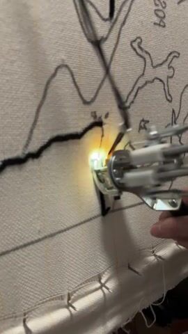 The video shows a close-up of a person's hand holding a tufting gun, which is being used to create a rug. The tufting gun is pressing against a canvas with a black outline of a design. The gun is actively tufting, with loops of black yarn emerging from the front of the tool and forming the design on the canvas. The canvas is stretched taut and appears to be a light beige color. The background is out of focus, but hints of the environment suggest an indoor setting. The lighting in the video is focused on the working area of the tufting gun, creating a bright glow. The person's fingers are visible holding the tufting gun, and the gun itself is a metal and plastic device with multiple tubes and a trigger mechanism. The overall impression is a snapshot of the rug-making process in action.