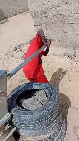 A person in a red outfit is pumping water from a hand pump into a black bucket. The hand pump is mounted on a stack of tires.  The person is standing on a sandy ground next to a gray concrete wall.  Water is flowing steadily from the pump into the bucket.  Other people are visible in the background.