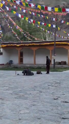 The video shows an outdoor scene at what appears to be a temple or monastery. In the foreground, there is a paved courtyard with two individuals. One person is dressed in dark clothing and is bowing down, appearing to be in a state of prayer or prostration. Another person, also dressed in dark clothing, stands nearby, observing. There are also bags on the ground. In the background, there is a building with a covered walkway supported by wooden pillars. Prayer flags are strung across the top of the building and extend into the distance. The environment appears to be mountainous.