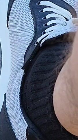 A close-up video of a person's foot in a white and black athletic shoe with white laces. The shoe is on a person's right foot, which is wearing a black sock. The person's leg and ankle are visible. The shoe is on a paved surface, possibly a sidewalk or patio. The background is blurred, but appears to be an outdoor setting with sunlight and shadows. The video is approximately 5 seconds long and is shot from a low angle.
