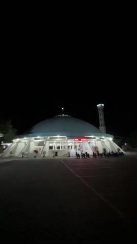 The video shows a mosque at night. The mosque has a large, dome-shaped roof that appears pale green. A tall minaret stands to the right of the mosque. Several motorcycles are parked in front of the mosque. The area in front of the mosque is paved with dark gray blocks. The sky is completely dark. The mosque is lit up by bright white lights along its edges and at the entrance, and a red sign above the entrance is visible. The camera is static throughout the video.