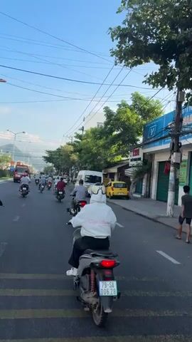 The video shows a street scene with several people riding motorbikes. The road is paved, and there are buildings and trees on both sides. The sky is blue with some clouds. The motorbikes are moving in the same direction, and some of them have passengers. The people on the motorbikes are wearing casual clothing. The scene appears to be in a city or town. The video also shows a white van and a red bus.