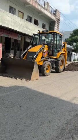 A yellow JCB backhoe loader is parked on a dirt road. A pile of dirt is next to it. Two people on a scooter ride past the backhoe. The scene appears to be in front of a building with a light pink facade. The sky is cloudy.