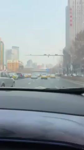 The view from the front seat of a car driving through a city street on an overcast day. The road is wide and has several lanes filled with cars, including many yellow taxis. On the right side of the road, pedestrians are waiting at a crosswalk. High-rise buildings line the street, and distant skyscrapers are visible in the background.