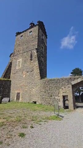 The video pans across a historical stone castle complex on a sunny day. On the left, a tall, square stone tower with small windows is visible. In the center, a stone archway leads through a thick stone wall. The wall appears aged and weathered, with some sections showing repairs. Beyond the archway, a paved path leads further into the castle grounds, with trees and another smaller tower with a green roof visible in the distance. The ground is a mix of gravel and grass, with some overgrown vegetation on top of the walls. A signpost is visible near the archway. The sky is bright blue with a single white cloud. The camera moves from left to right, offering a view of the castle walls and structures.