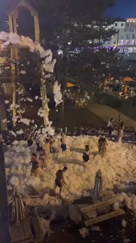A group of people, mostly young adults, are engaged in a foam party at night. They are dressed in swimwear and are interacting within a large pile of white foam. The foam appears to be dispensed from a structure on the left side of the frame. In the background, there are trees, lounge chairs, and a building with illuminated windows, suggesting an outdoor recreational area or resort. The lighting is dim, characteristic of nighttime, with artificial lights illuminating the scene. Some people are standing, while others are sitting or walking through the foam. The overall atmosphere seems to be one of fun and recreation.
