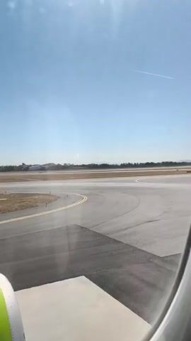 A plane is shown taking off from a runway, ascending into a clear blue sky. The sun is visible and bright, casting a light over the scene. The runway is paved and appears to be in good condition. The plane is seen from the perspective of another aircraft, as evidenced by the wing and window frame in the foreground. The other plane is positioned on the runway, possibly preparing for takeoff or taxiing. The environment is an airport with a flat landscape stretching out to the horizon.