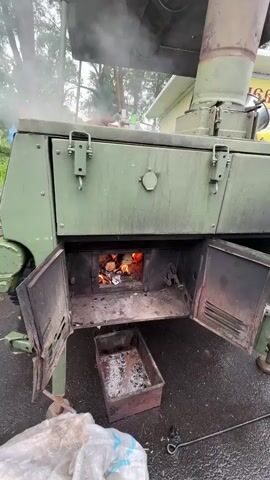 The video shows a close-up view of a military field kitchen, a large green metal oven with its firebox door open. Inside, a fire burns brightly with visible flames and glowing embers, with a few pieces of wood still burning. Ash and debris are collected in a metal tray beneath the firebox. Smoke billows out from the top of the oven. The oven itself appears to be mounted on a trailer, with a wheel visible on the left side and a support leg extended. The exterior of the oven is a dull green, showing signs of wear and tear.

In the background, partially visible through the smoke and out of focus, are trees and a light-colored building, suggesting an outdoor setting. The ground appears to be asphalt or a similar paved surface. A person wearing a camouflage uniform is visible on the far left, partially obscured by the oven. A white plastic bag lies on the ground in the foreground, with some dirt or debris spilling out.

The lighting and the visible smoke suggest that the activity is taking place during the day, possibly in overcast or early morning conditions, as there are no harsh shadows. The overall scene evokes a sense of practical, utilitarian activity, likely related to preparing food in a field setting. The "Местечко Раифа, Russia" context indicates the geographical location of this scene. There is no visible text in the video.