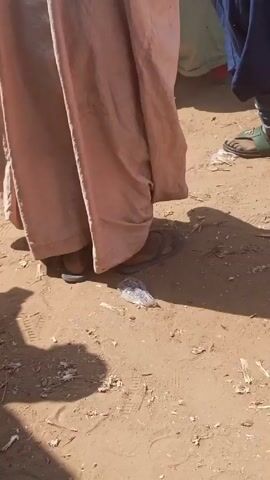 The video shows the lower portion of a person wearing a long, light brown robe and dark sandals. They are standing on a dirt ground. To the right of the person, another individual is partially visible, wearing a blue garment and green sandals. A piece of clear plastic litter is on the ground near the feet of the person in the brown robe. The background is blurry and suggests other people are present. The ground is dry and dusty. There are shadows cast on the ground.