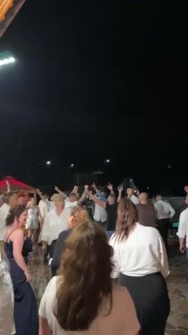 A group of people dance at night. Many people, mostly adults, are gathered outdoors, dancing to music. The camera is positioned behind some of the dancers, looking forward at the rest of the group. Some people are raising their hands in the air, while others are swaying and moving to the beat. The setting appears to be an outdoor event, possibly a party or celebration, with some lights visible in the background illuminating the space. Some people are wearing white shirts. One person in the background on the right is holding a bottle up in the air.