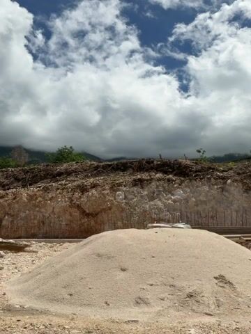This 10-second video shows an outdoor construction or excavation site in Bratešići, Montenegro, on a partly cloudy day. The foreground features a large pile of light-colored rocks or debris and a mound of sand. To the left, a utility pole is partially visible. The central focus is a significant cut-away rock face, revealing layers of earth and rock. This rock face appears to be reinforced with vertical rebar or mesh embedded within it. Above the rock face, a dense line of green trees and brown earth indicates the original ground level. The sky is a mix of bright blue patches and large, fluffy white clouds. No people are visible, and no specific activity is taking place; it's a static shot of the landscape. Some faint red markings, possibly spray paint, are visible on the rock face but are not legible.
