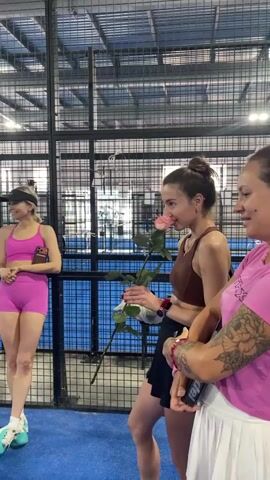 Three women are standing in an indoor paddle court. One woman on the left, wearing a pink sports bra and pink shorts, is looking to her right. The woman in the middle is wearing a brown sports bra and a black skirt, holding a long-stemmed pink rose. The woman on the right, wearing a pink shirt, has tattoos on her arm and is looking at the woman with the rose. The court has a blue surface and black net fencing. The woman in the middle appears to be smelling the rose. Another woman in a white long-sleeved top and black shorts is in the background, on the left side of the court.
