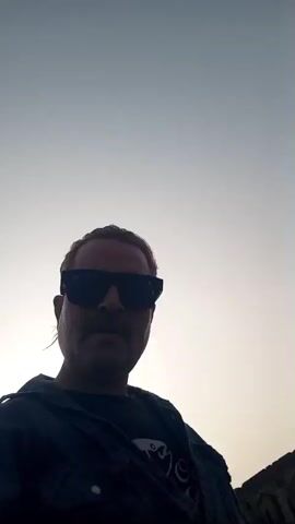 The video shows a man in sunglasses, a dark t-shirt, and a plaid shirt taking a selfie. He appears to be outdoors in a dusty or sandy area. Behind him, there are historical-looking stone structures with domed roofs. In some shots, a road and some greenery are visible. There are motorcycles in the background in one of the shots. The sky is bright and appears to be a clear day. The camera angle is from a low perspective, looking up at the man's face. The man is standing relatively still, taking a video selfie.