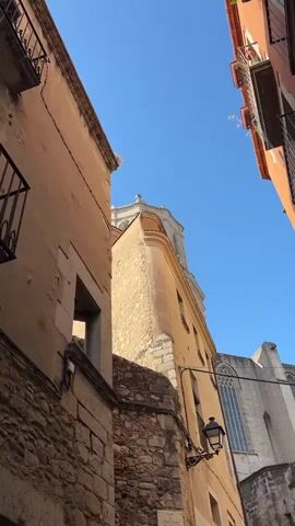 The video shows a low-angle view of buildings and a church tower against a clear blue sky. The buildings on the left and right are close together, forming a narrow alleyway. The building on the left is made of stone and stucco, with a balcony. The building in the center is yellow, with a stone foundation. The church tower, topped with a dome and cross, is visible above the buildings. A large gothic window is visible on the right side of the frame. The scene is illuminated by sunlight. There are no people visible in the video.