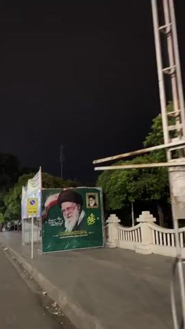 The video shows a large green banner with a portrait of a man with a white beard and glasses, wearing a brown hat. The banner also features a smaller portrait in the upper right corner. There is text in a language that appears to be Arabic or Persian on the banner. To the left of the banner is a yellow sign with a blue circle and a symbol that may indicate a taxi stand. There are also flags and other banners visible in the background. The scene is outdoors at night, with trees and a white railing visible on the right side. The ground is paved and appears to be a sidewalk or street.